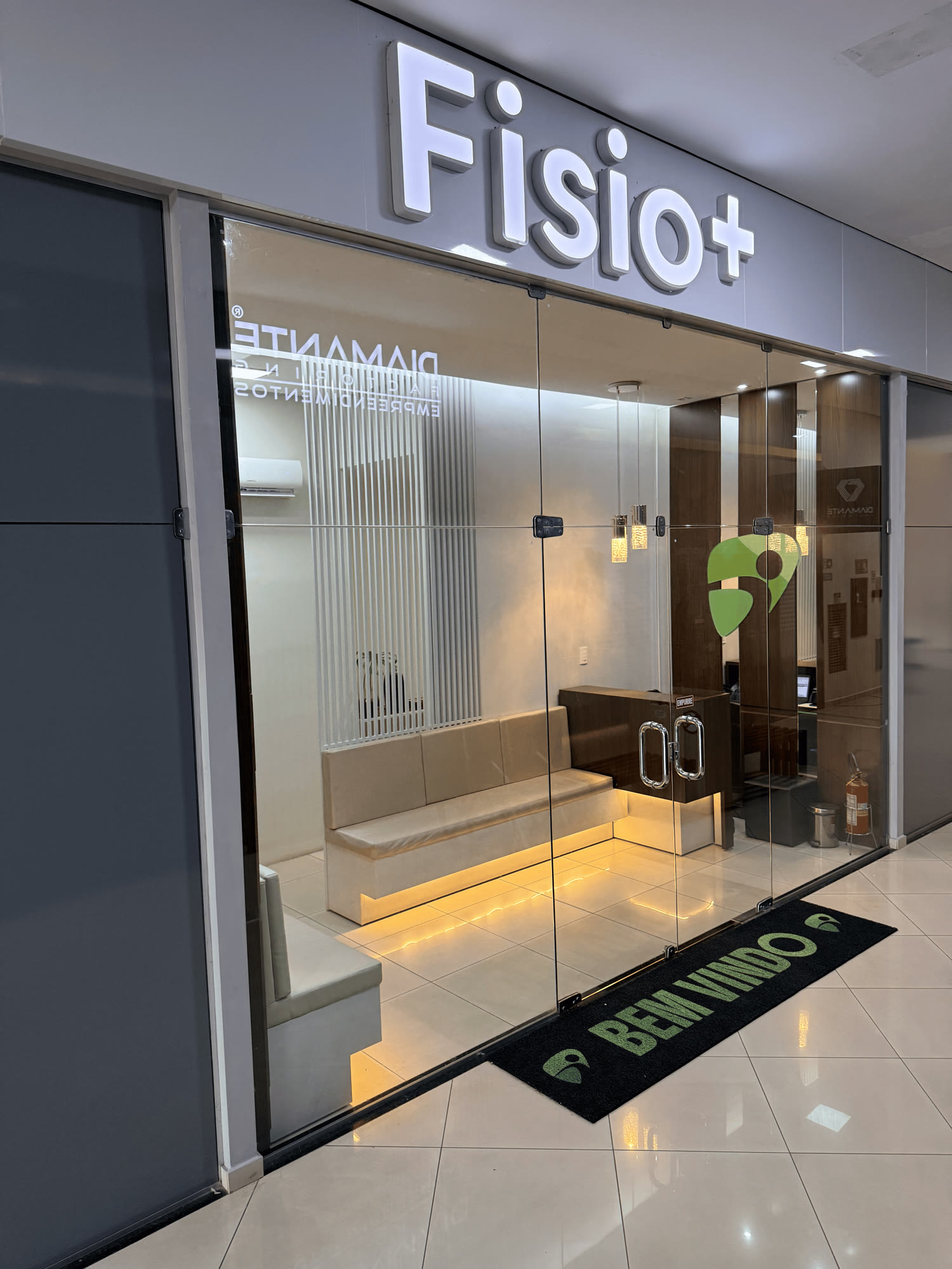 Fachada da Clínica Fisio+, com letreiro iluminado e tapete de boas-vindas na entrada